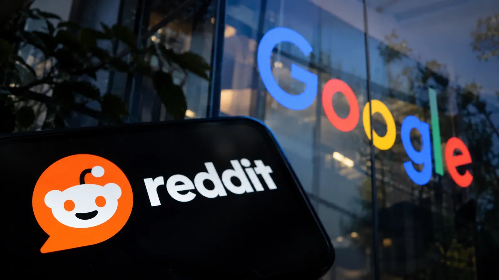 Reddit logo on a phone screen in front of a Google sign on a glass building