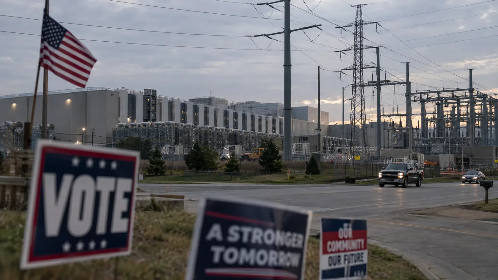 Election yard signs reading VOTE and A Stronger Tomorrow in the foreground, with a large data center and power transmission towers at dusk behind them